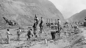 Panama Canal construction in 1913 showing workers drilling holes for dynamite in bedrock, as they cut through the mountains of the Isthmus. Steam shovels in the background move the rubble to railroad cars.