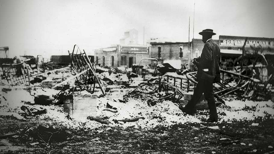 A Black man with a camera looks at the skeletons of iron beds which rise above the ashes of a burned-out block after the massacre in June 1921.