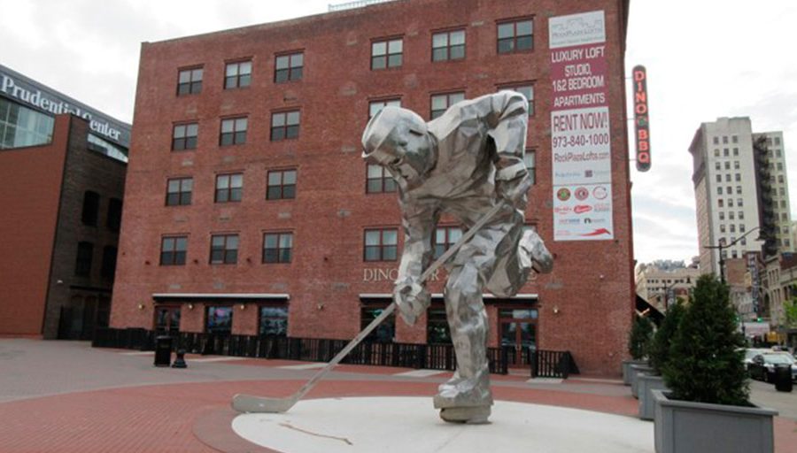 "The Iron Man" sculpture of a hockey player next to the Prudential Center in Newark, where the New Jersey Devils hockey team plays. In the background, signs can be seen for Dinosaur Bar-B-Cue BBQ and Rock Plaza Lofts luxury apartments.