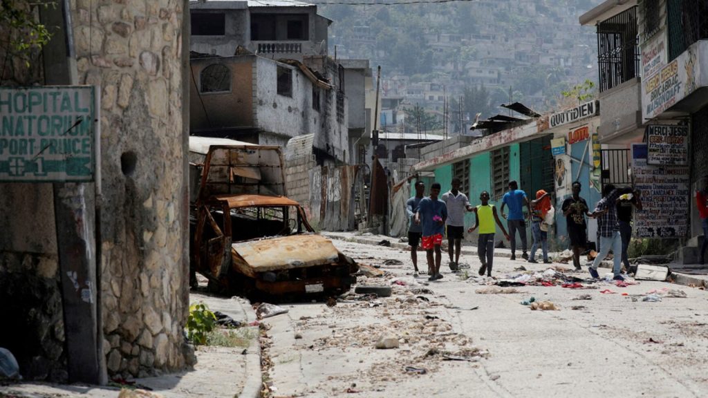 People walk past a damaged car in the Carrefour Feuilles neighborhood, which was deserted due to gang violence, in Port-au-Prince, Haiti March 19, 2024. REUTERS/Ralph Tedy Erol