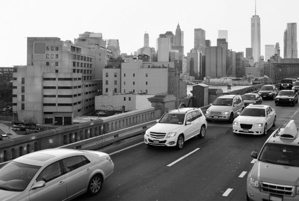 This image captures heavy traffic during sunrise or sunset on the Brooklyn Bridge, with the Manhattan skyline prominently featuring the One World Trade Center in the background.